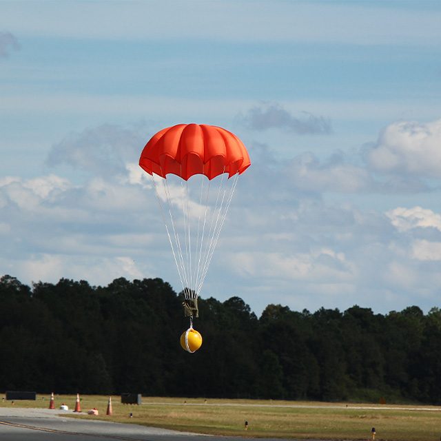 A large yellow weight lands beneath an orange round parachute during a test jump for research and product development with Mobility Lab Inc